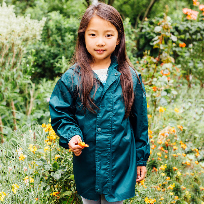 A young girl in a teal raincoat stands amongst flowers. She holds a small orange flower in her hand.